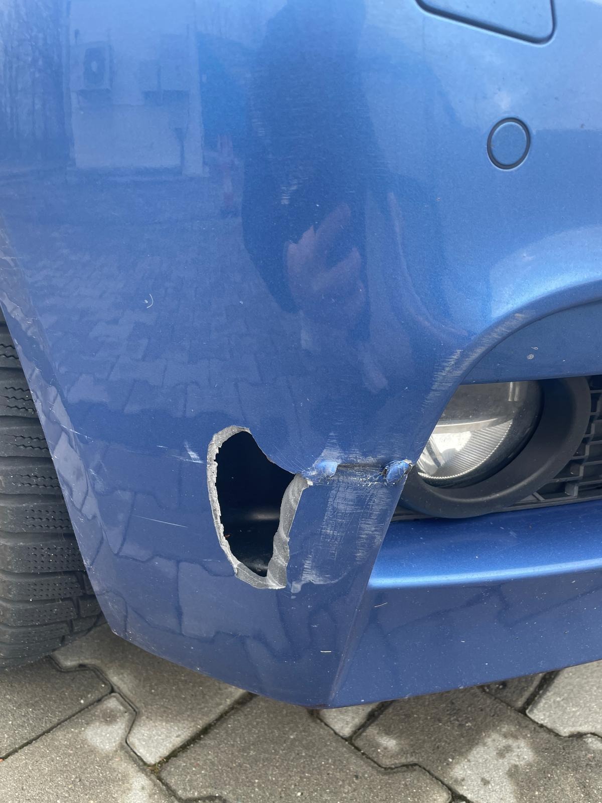 Uszkodzony, niebieski przedni zderzak samochodu z widoczną dziurą i zarysowaniami, opona w tle.