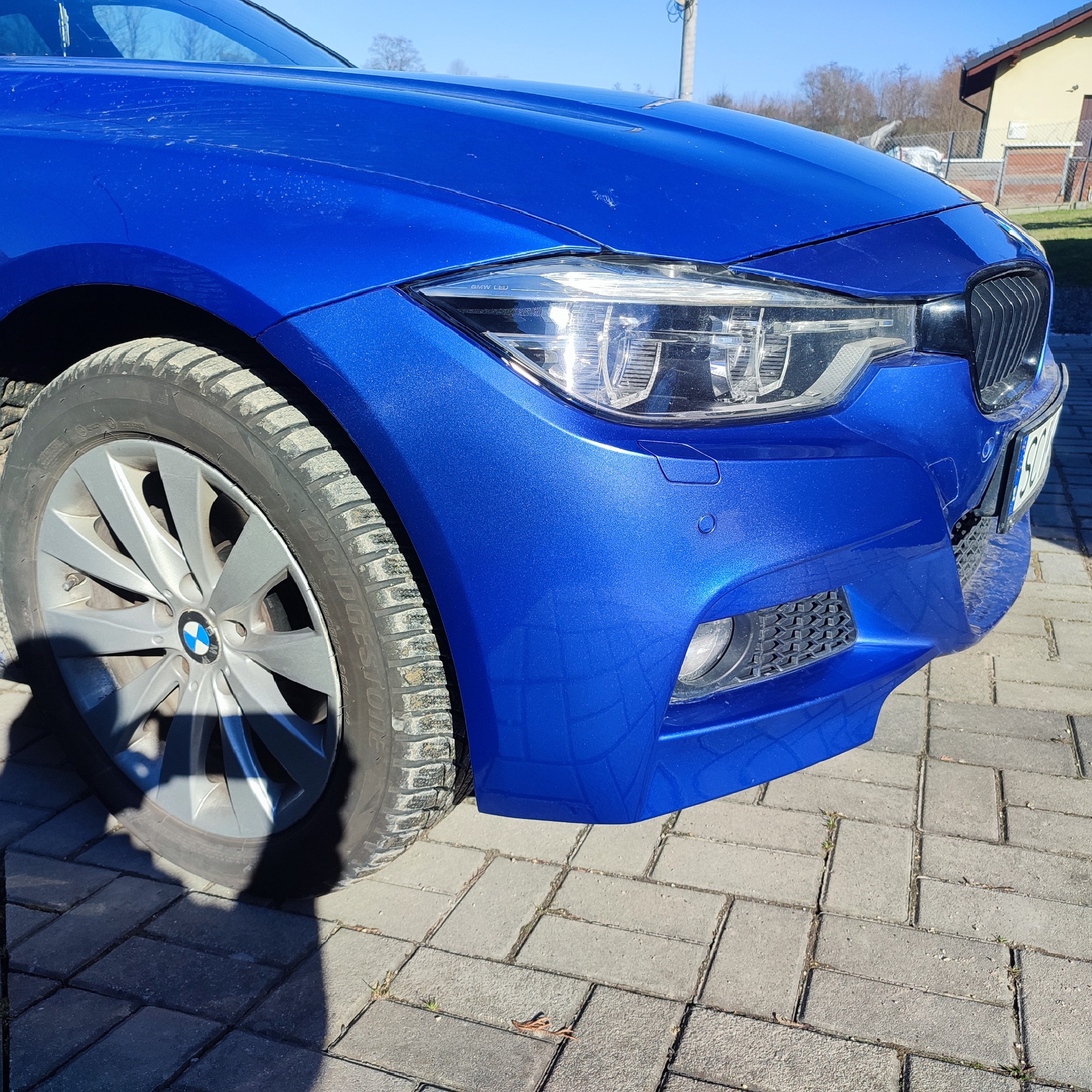 Niebieski samochód BMW, widoczny fragment przodu z reflektorem, kołem i błotnikiem, zaparkowany na szarej kostce brukowej.