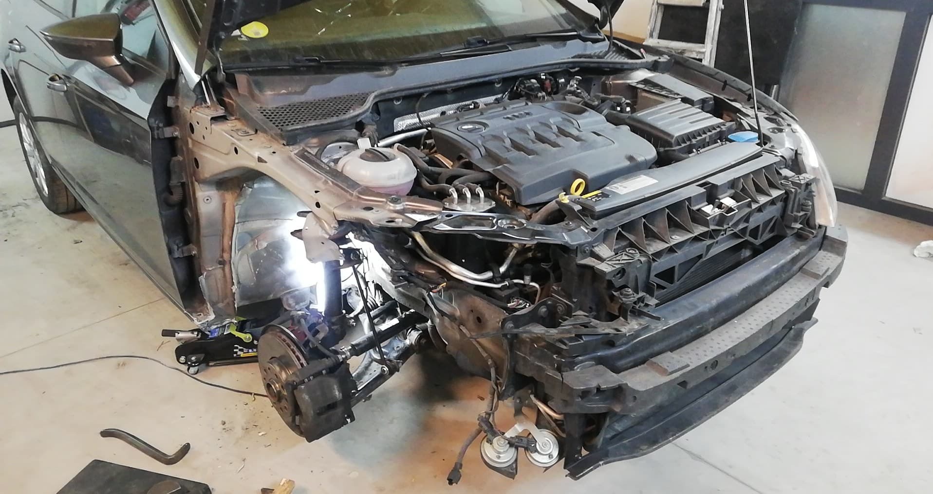 Samochód osobowy w trakcie naprawy karoserii, widoczny otwarty przedział silnika, brak przedniego zderzaka, reflektorów i błotnika po stronie kierowcy, lampa warsztatowa oświetla nadkole.