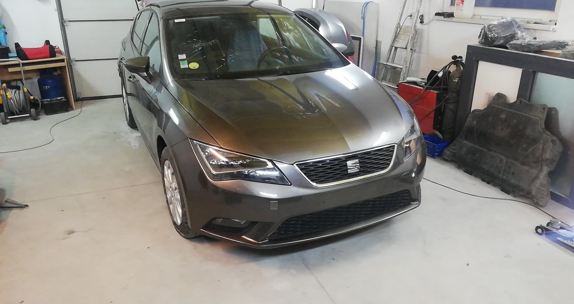 Brązowy samochód marki Seat Leon po naprawie karoserii, zaparkowany w warsztacie. Na przedniej szybie widoczne ślady po naprawie.