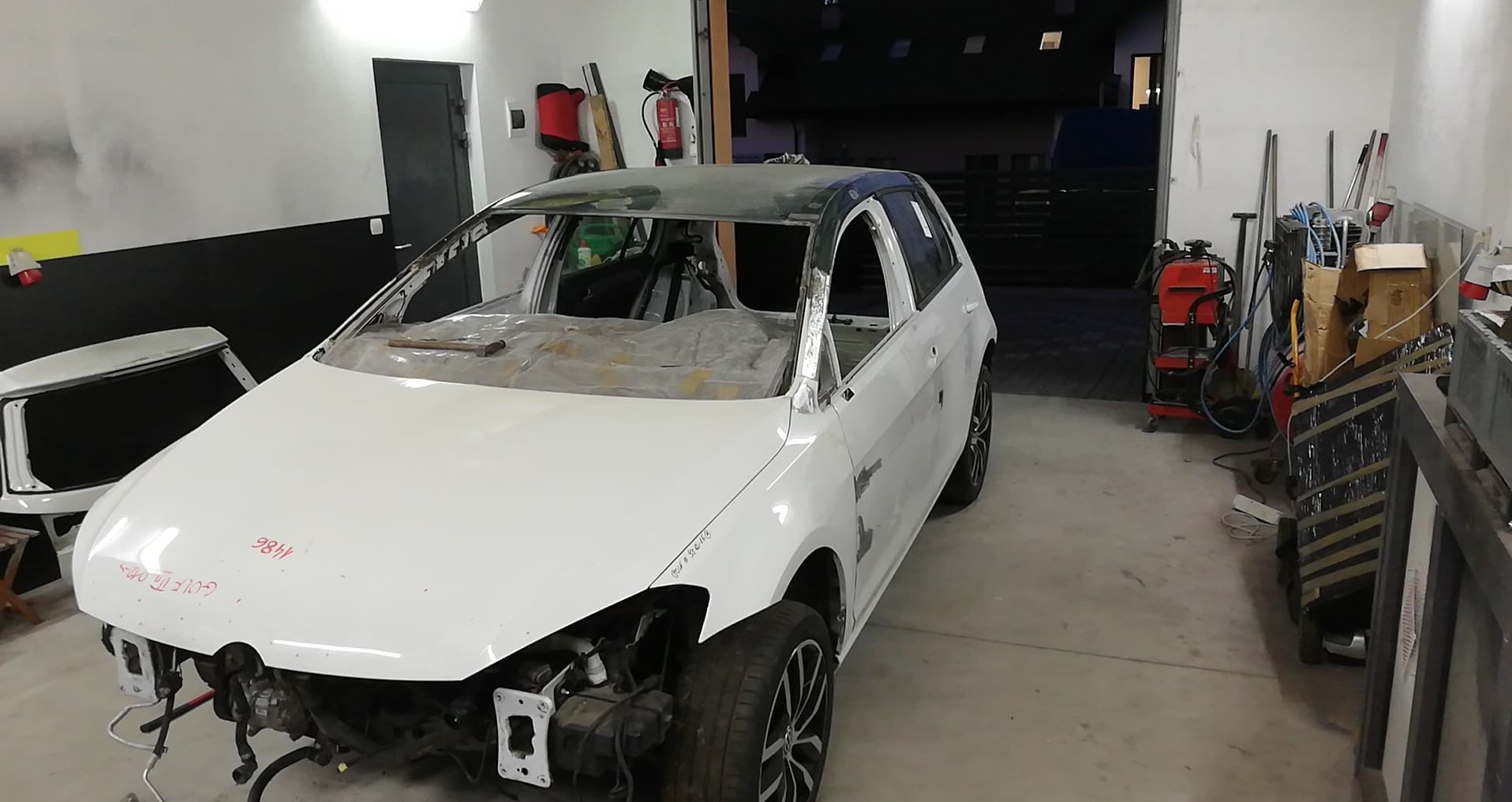 Biały samochód marki Golf bez szyb i drzwi, przygotowany do lakierowania w warsztacie, z widocznymi śladami szpachlowania i notatkami na karoserii.