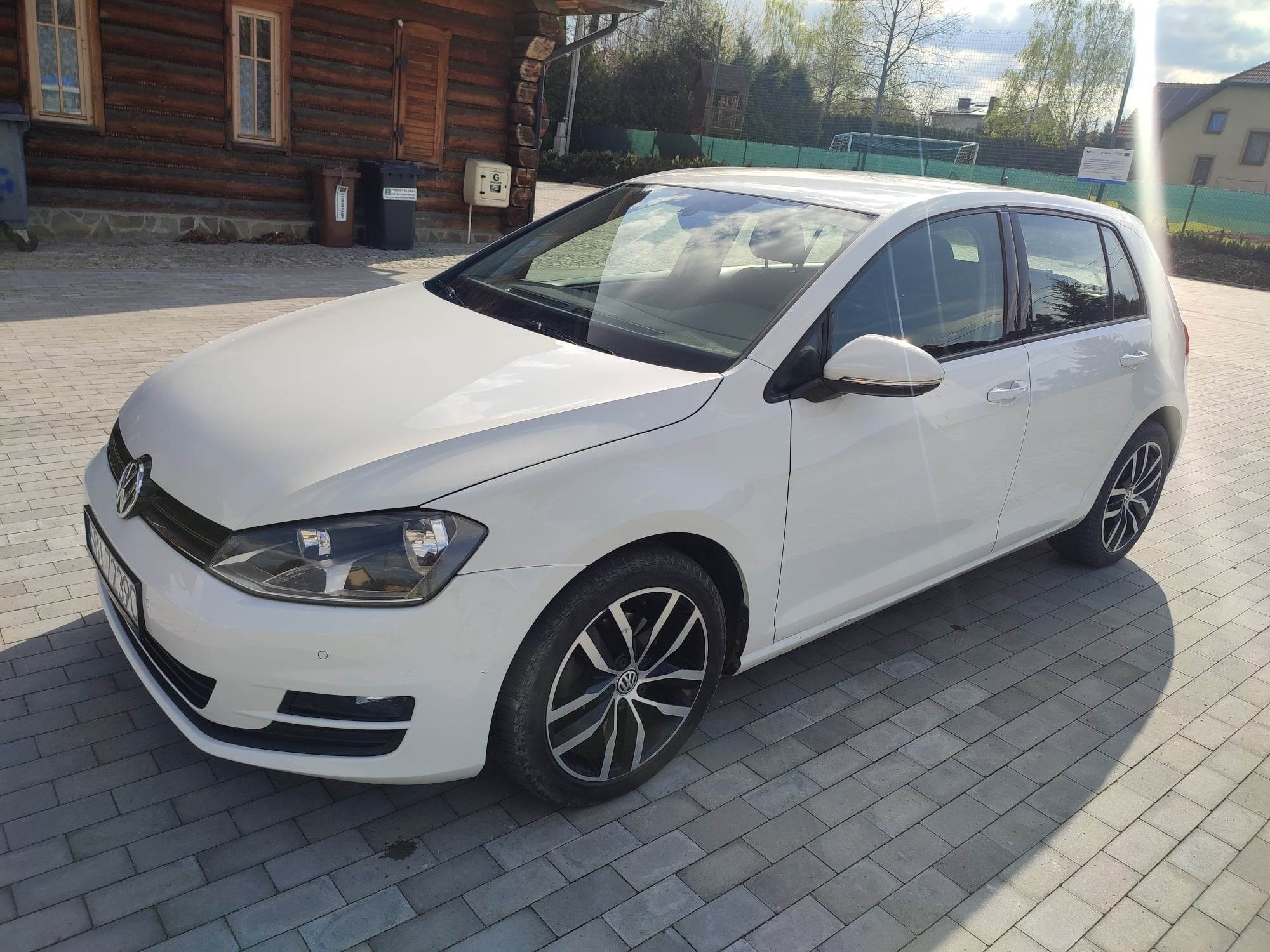 Biały samochód marki Volkswagen Golf zaparkowany na szarym bruku, z drewnianym budynkiem w tle.
