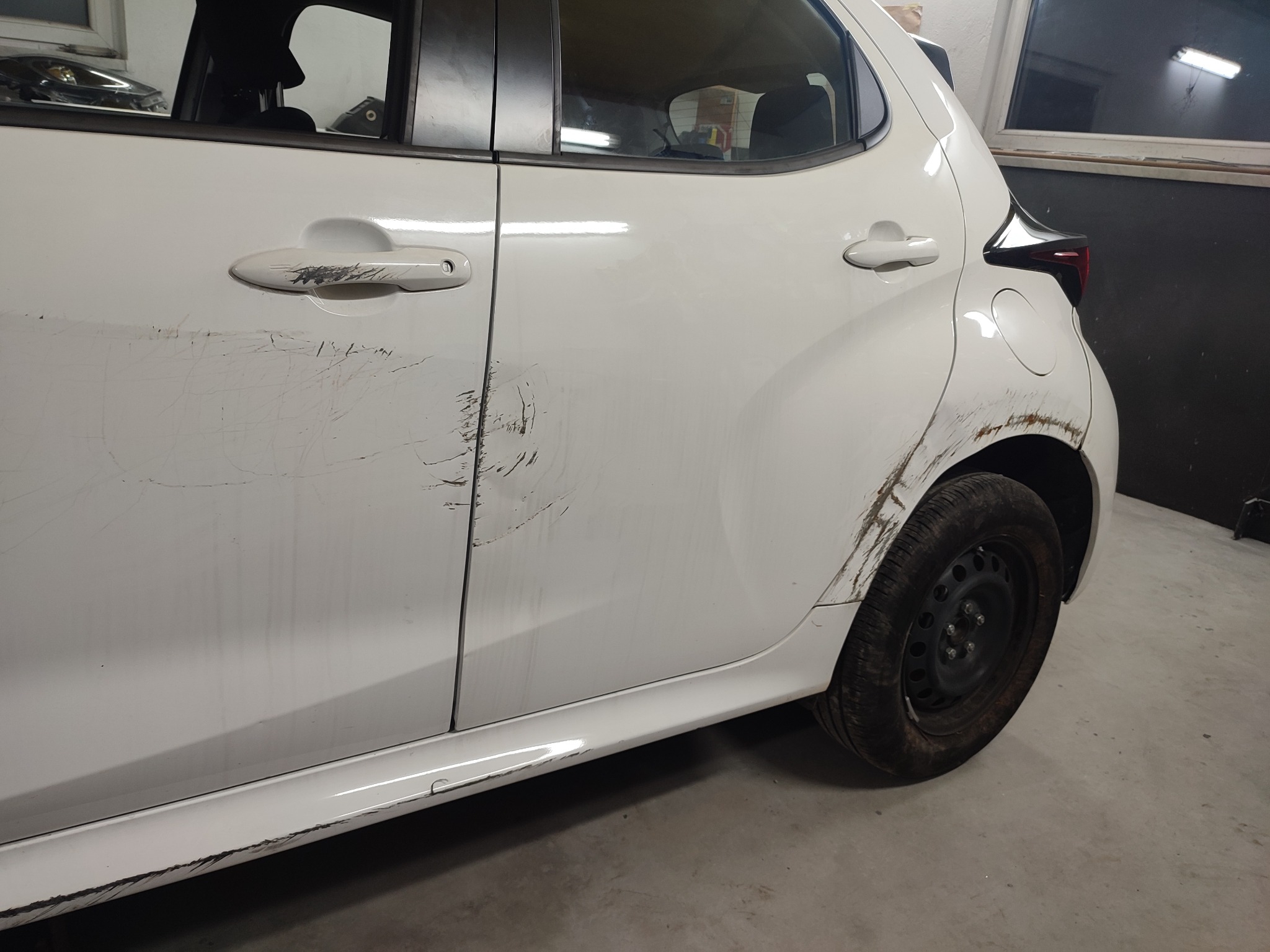 Biały samochód z widocznymi uszkodzeniami karoserii (rysy, otarcia) na drzwiach i błotniku, sfotografowany w warsztacie w Międzyrzeczu Górnym przed naprawą.