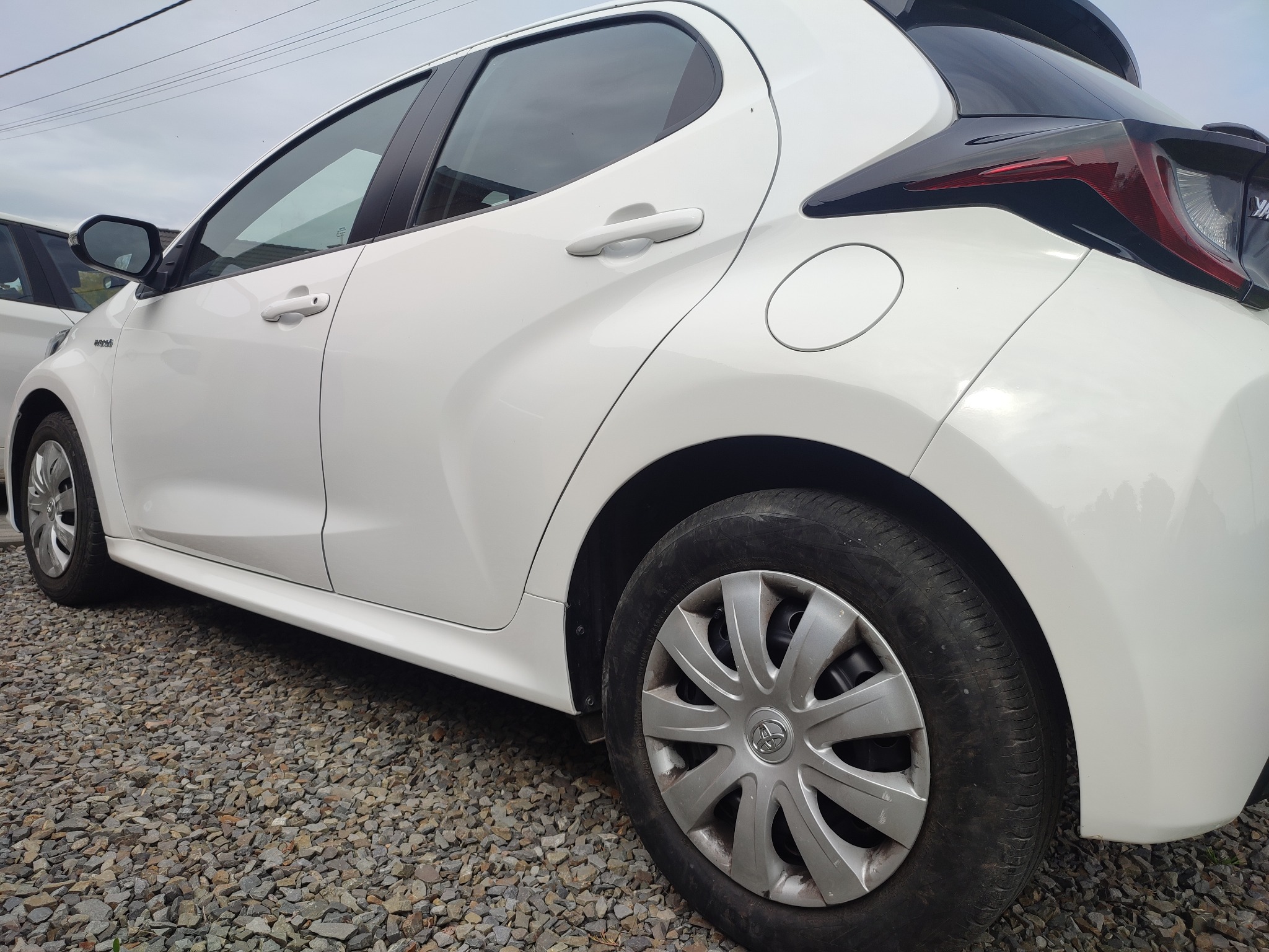 Biały samochód osobowy, widok z boku, na żwirowym podłożu, z widocznym tylnym kołem i fragmentem karoserii.