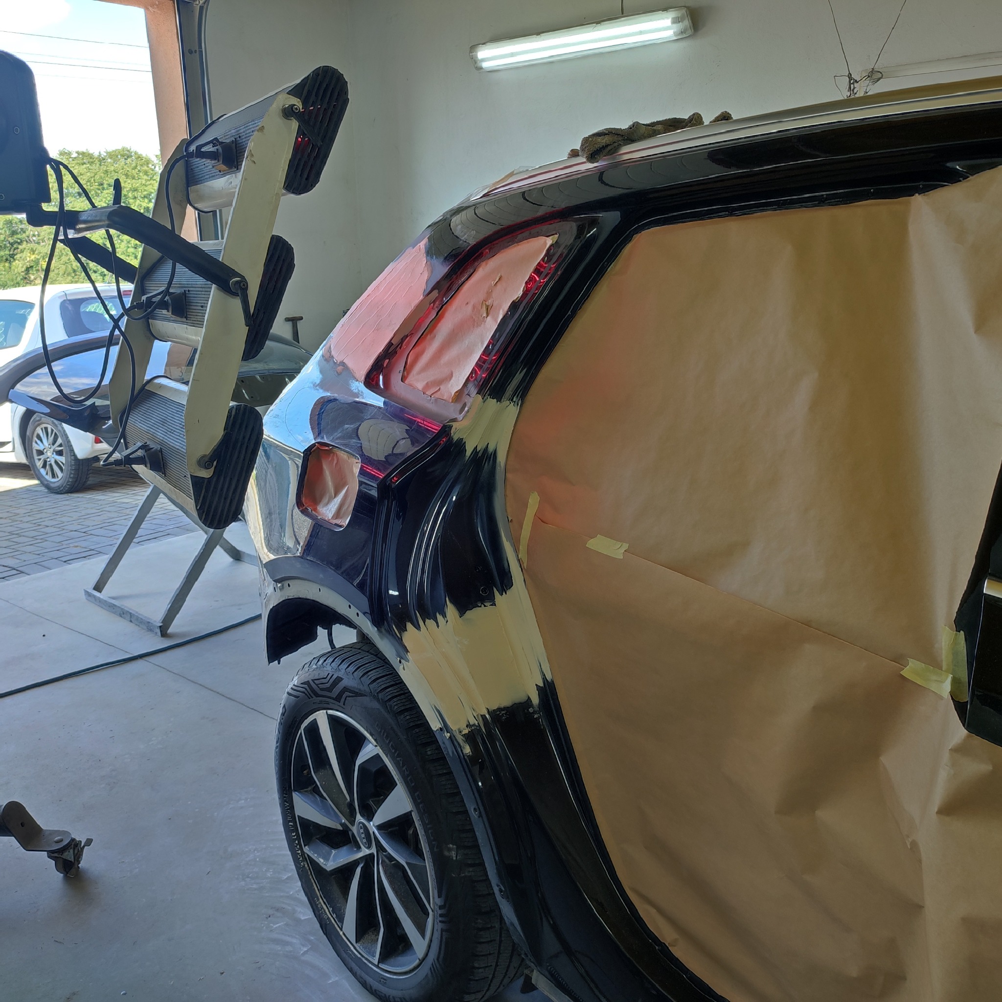 Czarny samochód osobowy w trakcie naprawy karoserii, zabezpieczony papierem maskującym, z widocznym urządzeniem do suszenia lakieru i szpachlą na błotniku.