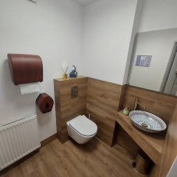 Remonty-Wykończenia wnętrz Łukasz Pajdowski - Nowoczesna łazienka z drewnopodobnymi płytkami, dekoracyjną umywalką i minimalistyczną toaletą podwieszaną. Uwagę zwraca elegancki dozownik na papier i dekoracje na zabudowie.