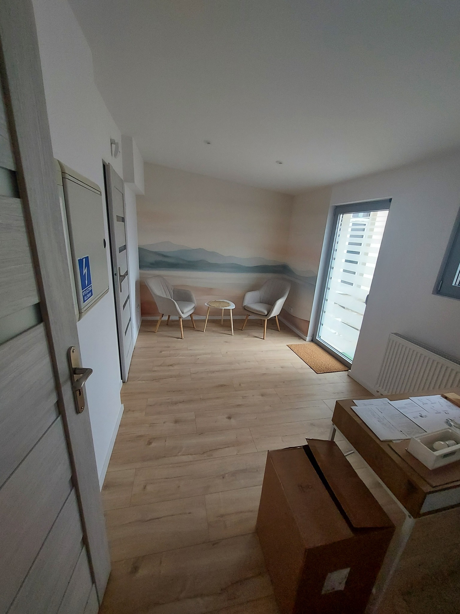 Jasne wnętrze po wykończeniu: drewniana podłoga, ściana z motywem górskim, okno z żaluzjami. Widoczne krzesła i stolik tworzą przytulną strefę relaksu.