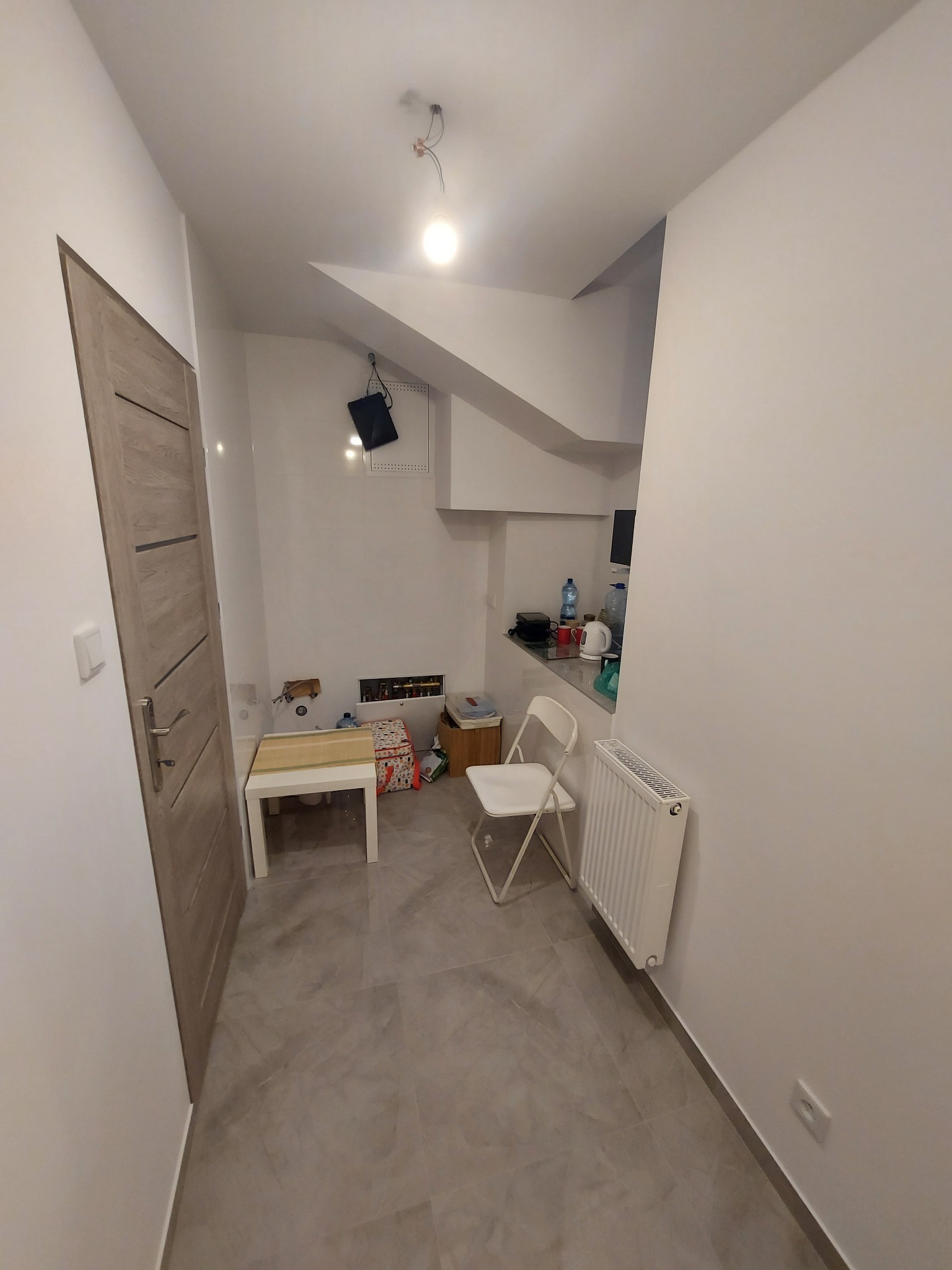 Wnętrze korytarza z szarą podłogą, białe ściany i drewniane drzwi. Widoczny grzejnik, krzesło i mały stolik. Częściowo widoczna kuchnia w tle.