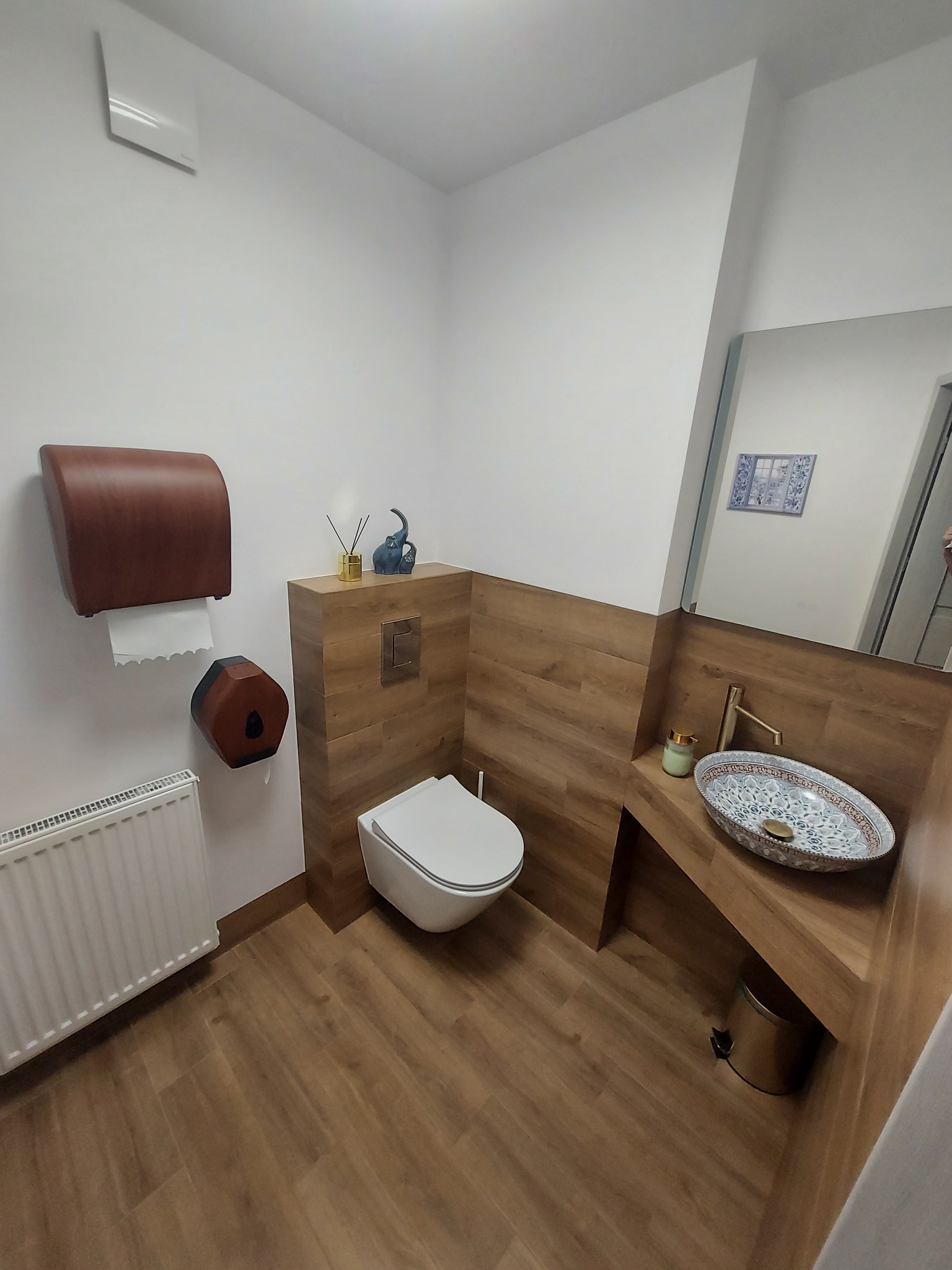 Nowoczesna łazienka z drewnopodobnymi płytkami, dekoracyjną umywalką i minimalistyczną toaletą podwieszaną. Uwagę zwraca elegancki dozownik na papier i dekoracje na zabudowie.