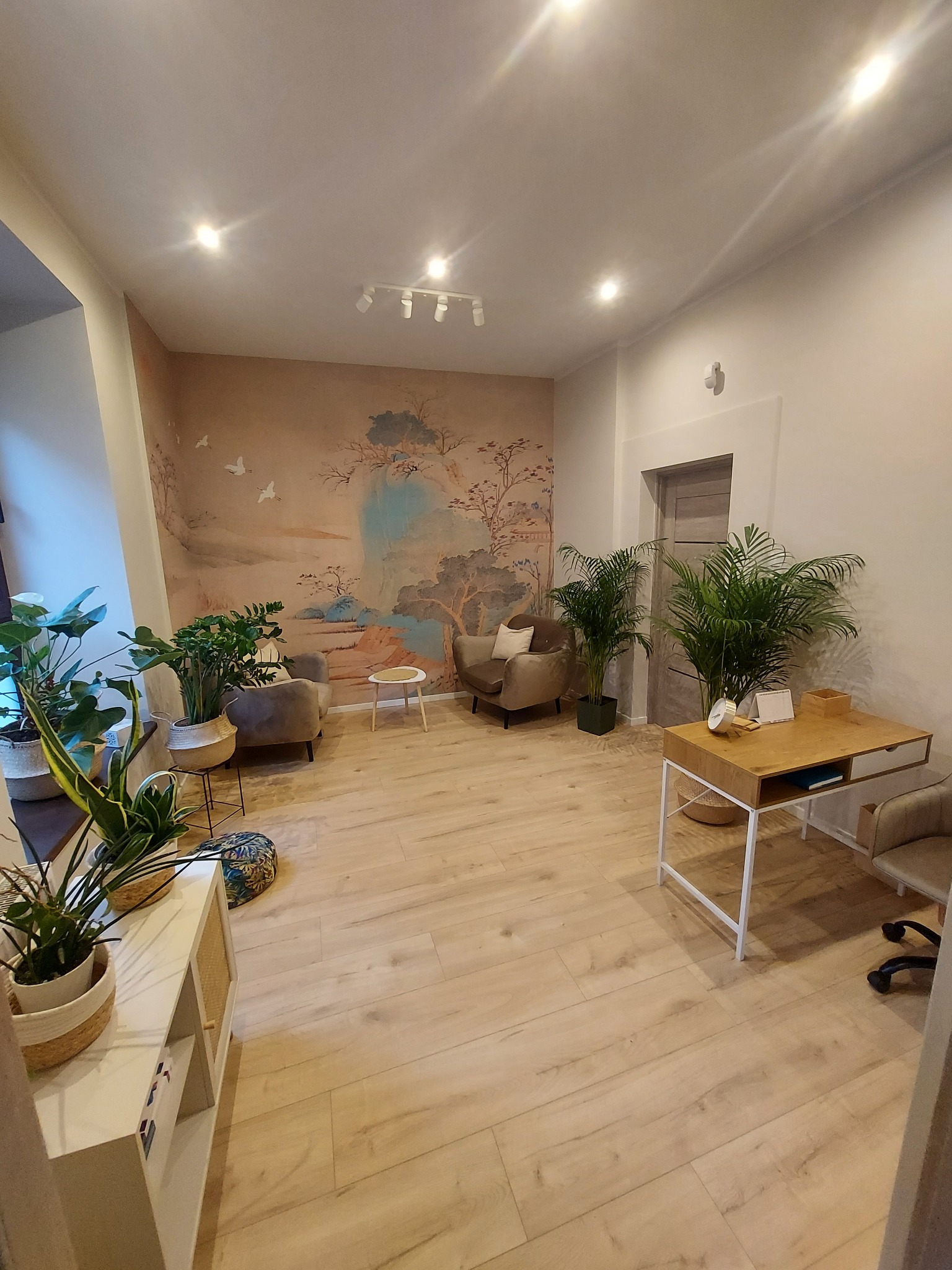 Jasne wnętrze z fototapetą w stylu orientalnym, drewnianą podłogą i zielonymi roślinami doniczkowymi. Dwa fotele i biurko tworzą przytulną przestrzeń.