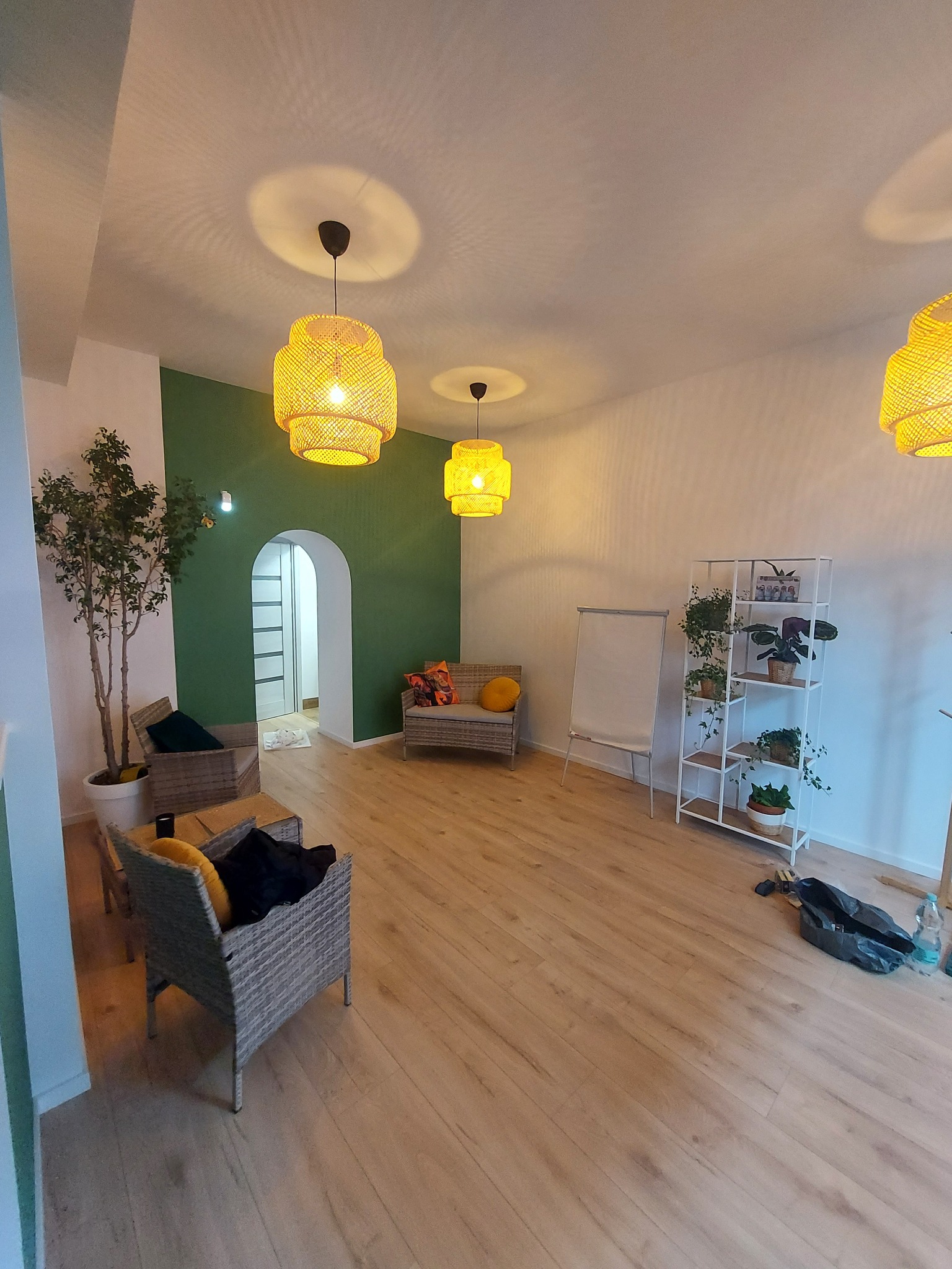 Jasne wnętrze po remoncie: drewniana podłoga, zielona ściana z łukiem, rattanowe meble i lampy. Nowoczesny design z roślinami doniczkowymi.