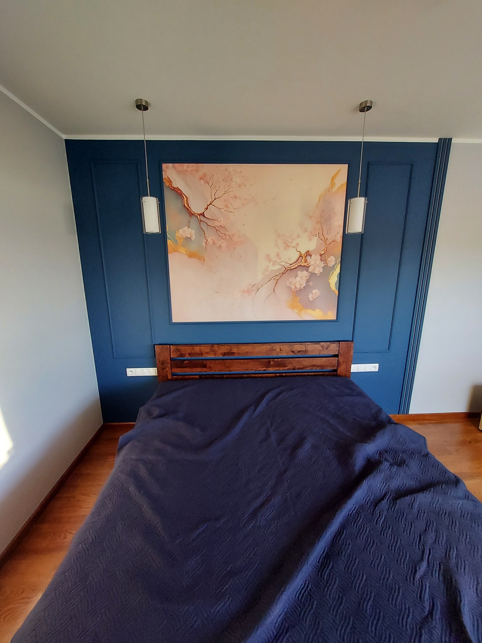 Sypialnia z drewnianym łóżkiem i niebieską ścianą z dekoracyjnymi listwami. Nad łóżkiem wisi obraz z motywem kwitnących gałęzi i dwie wiszące lampy.