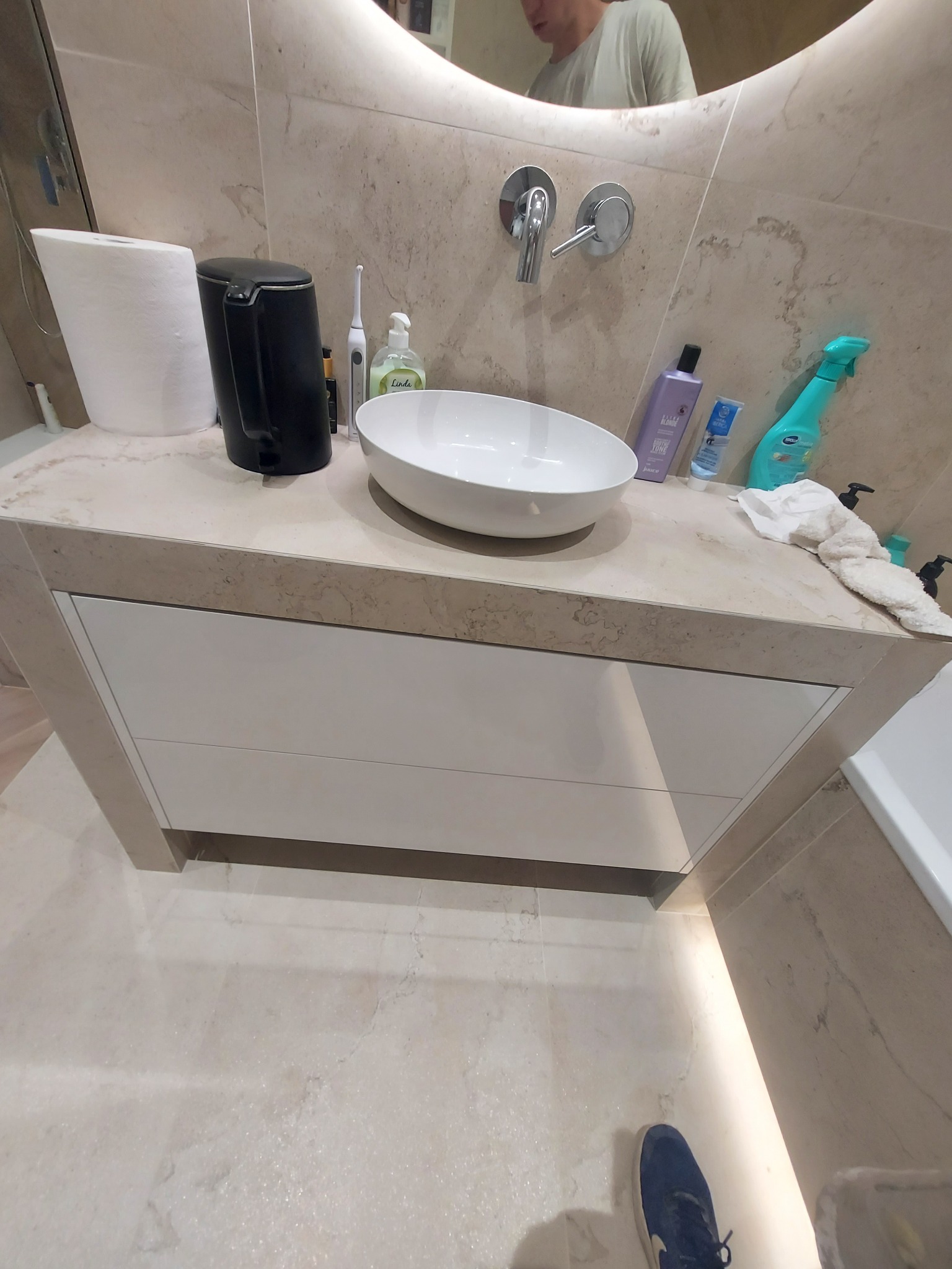 Beżowa łazienka z owalną umywalką nablatową, armaturą i akcesoriami. Widoczne odbicie osoby w lustrze. Blat z imitacją marmuru i białe szafki pod umywalką.