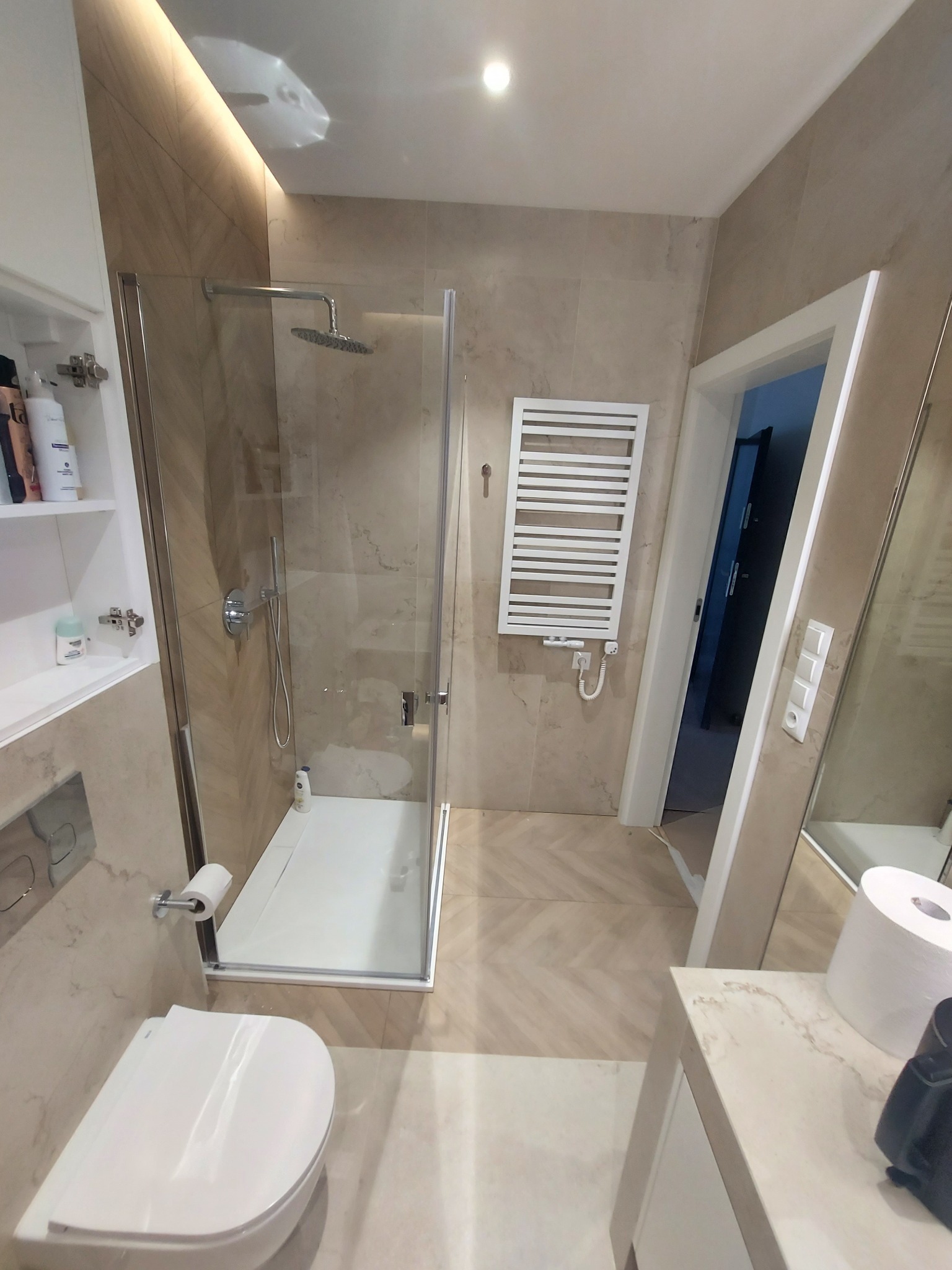 Nowoczesna łazienka po remoncie: kabina prysznicowa ze szklanymi drzwiami, podwieszana toaleta, grzejnik drabinkowy i płytki imitujące kamień.