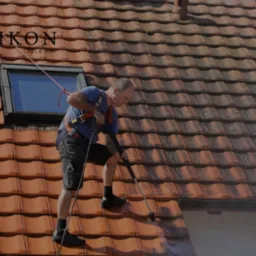Pracownik w uprzęży myje dachówkę na dachu domu za pomocą myjki ciśnieniowej. Część dachu jest już umyta. Widoczne okno dachowe i logo firmy na górze.
