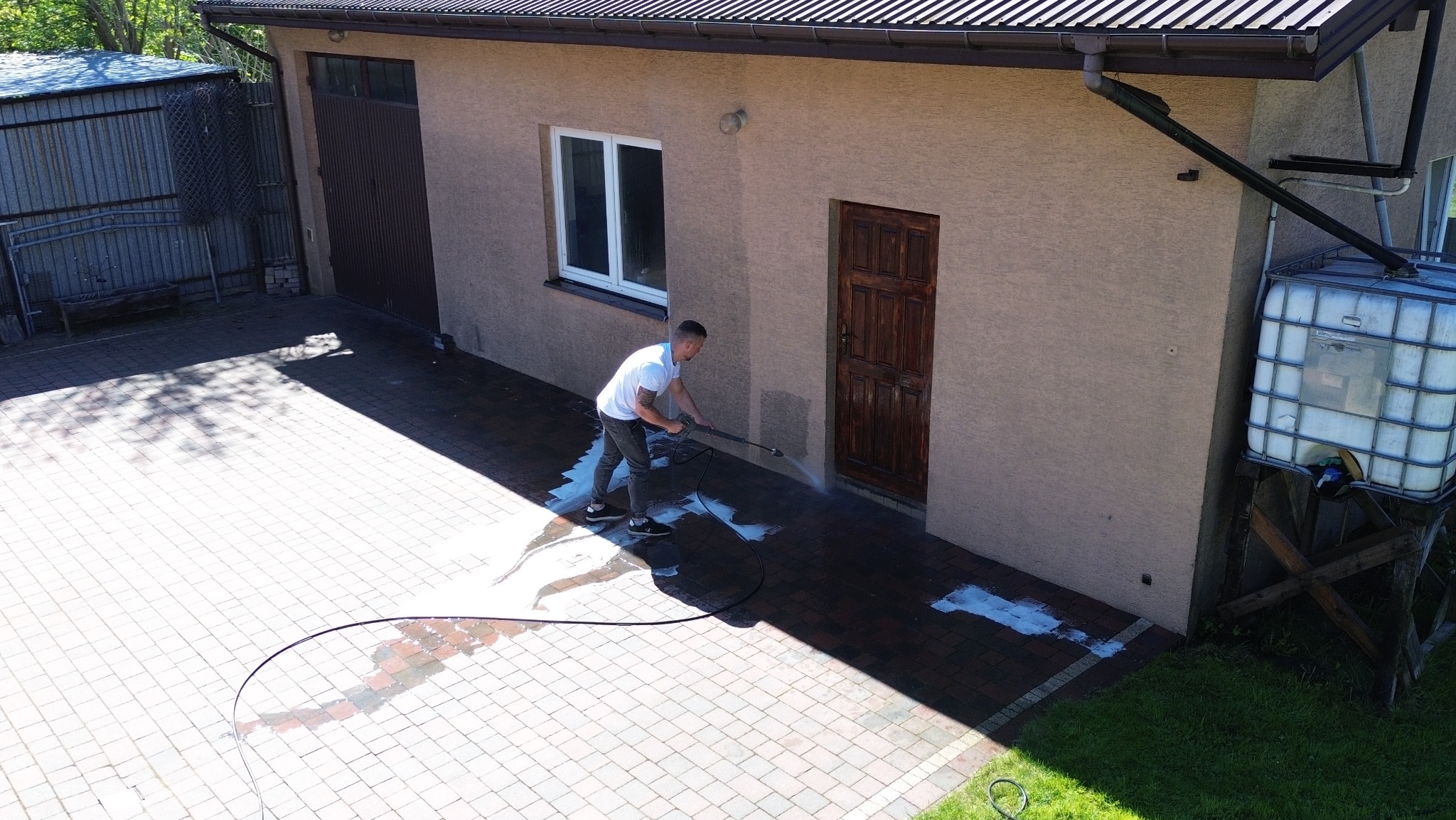 Mężczyzna czyści kostkę brukową myjką ciśnieniową na podjeździe przed budynkiem z beżową elewacją i brązowymi drzwiami. Widoczny zbiornik na wodę.