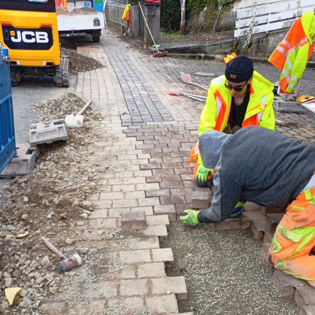 Układanie kostki brukowej na posesji. Dwóch robotników w odblaskowych ubraniach pracuje przy układaniu nowej nawierzchni z kostki. W tle widoczny sprzęt budowlany JCB.