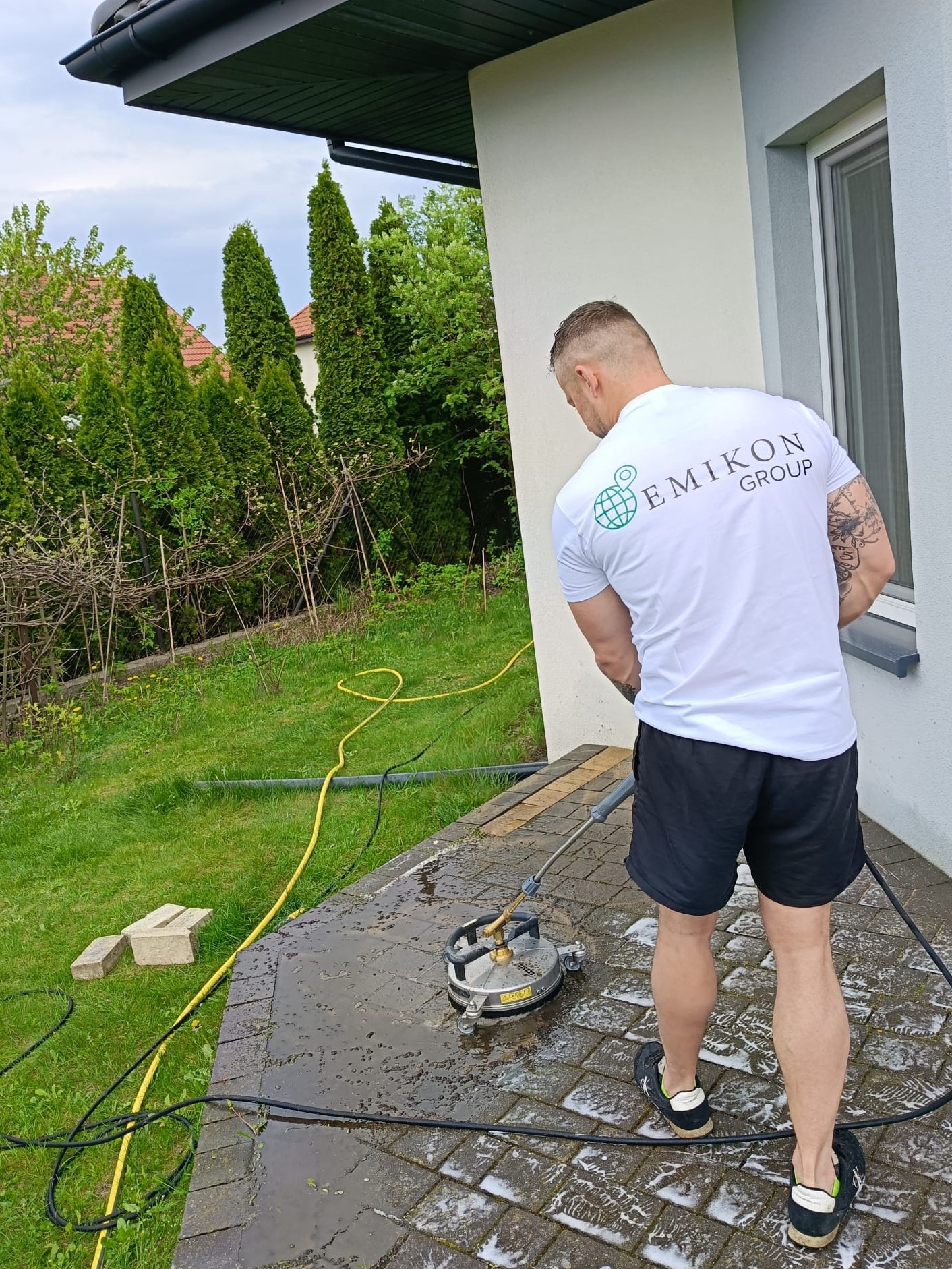 Mężczyzna w koszulce firmy Emikon Group czyści kostkę brukową obrotową myjką ciśnieniową na zewnątrz budynku. Widoczny żółty wąż i trawnik w tle.
