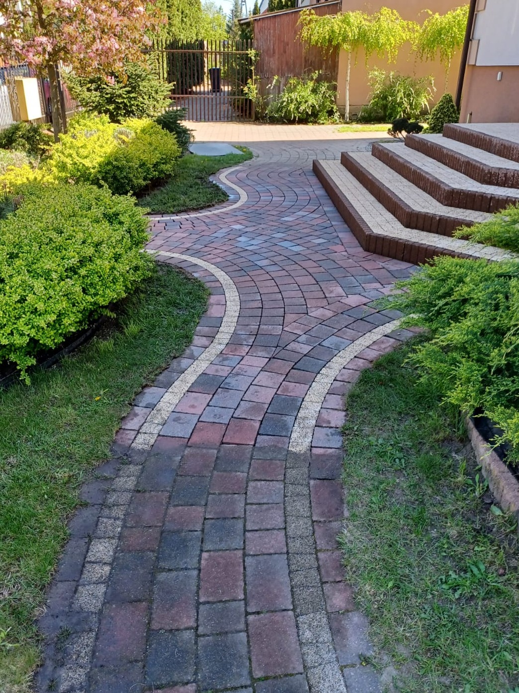 Kręta ścieżka z kostki brukowej w odcieniach czerwieni i szarości, obramowana jasnym żwirem, prowadząca do schodów wejściowych. Otoczenie zdobi trawa i krzewy.