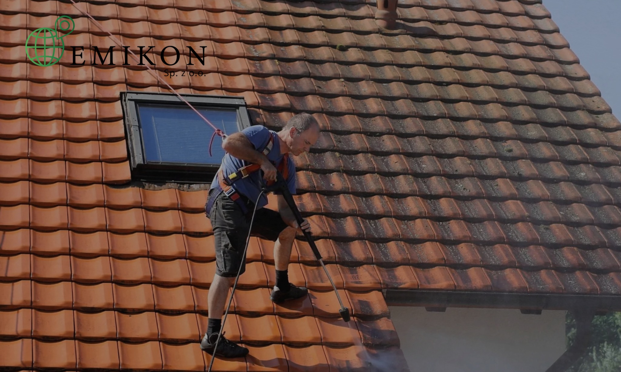 Pracownik w uprzęży myje dachówkę na dachu domu za pomocą myjki ciśnieniowej. Część dachu jest już umyta. Widoczne okno dachowe i logo firmy na górze.