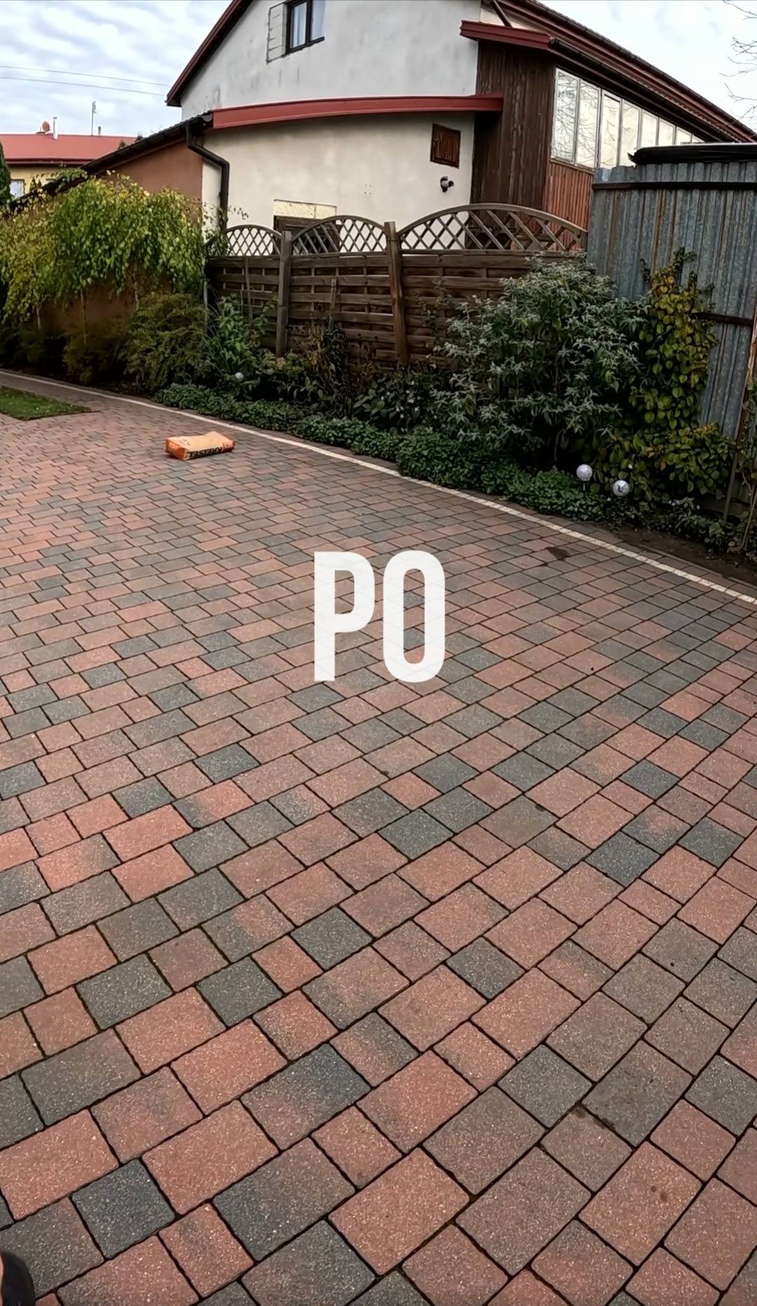 Czysta kostka brukowa po umyciu, widok z góry, zarys budynku w tle. Kostka w odcieniach czerwieni i szarości, zarysowana białą linią oddzielającą od trawnika.