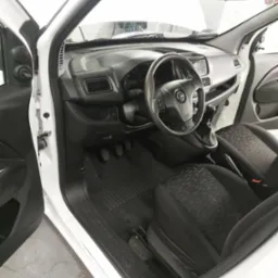 Wnętrze białego samochodu dostawczego z czarną tapicerką foteli i gumową wycieraczką pod nogami, widoczne pedały, kierownica z logo, otwarte drzwi kierowcy.