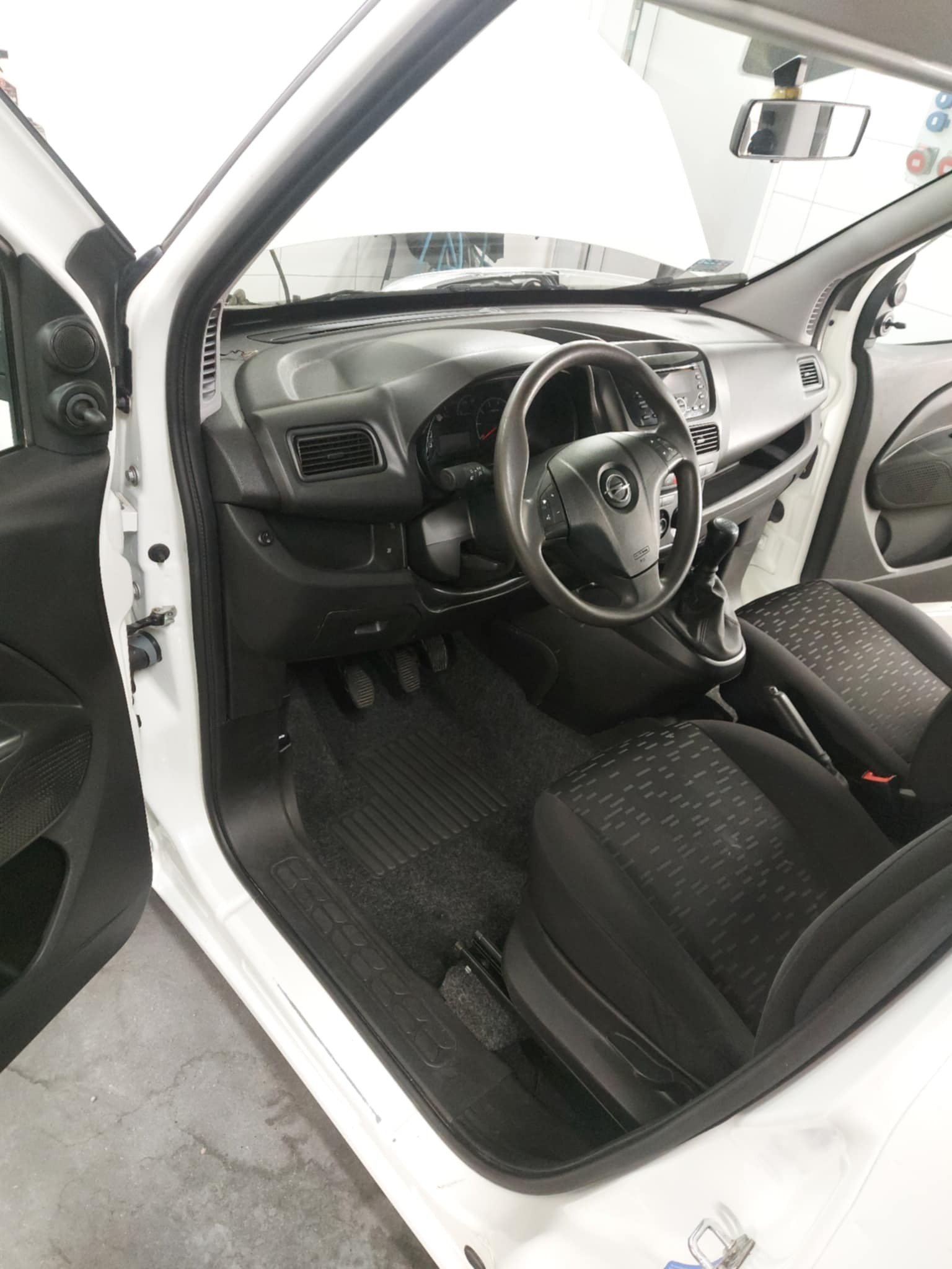 Wnętrze białego samochodu dostawczego z czarną tapicerką foteli i gumową wycieraczką pod nogami, widoczne pedały, kierownica z logo, otwarte drzwi kierowcy.