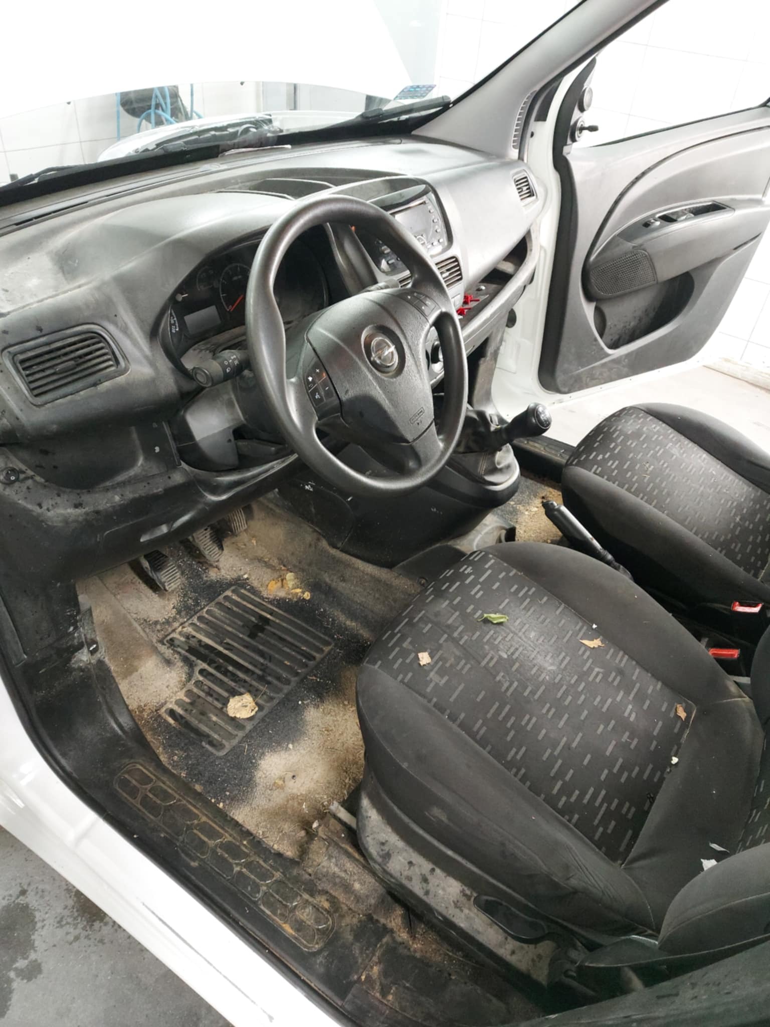 Wnętrze bardzo brudnego samochodu z widocznymi plamami i zabrudzeniami na fotelach, podłodze i desce rozdzielczej. Widoczne liście i inne zanieczyszczenia na podłodze.