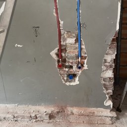 Hydraulika u Bartka - Wykute w ścianie bruzdy z wystającymi rurami instalacji wodnej (czerwona i niebieska) w trakcie montażu, widoczne fragmenty cegieł i ślady po kuciu.