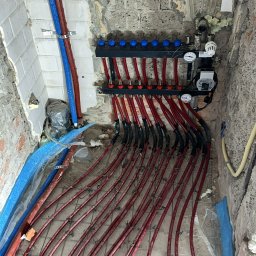 Hydraulika u Bartka - Rozdzielacz ogrzewania podłogowego z czerwonymi rurami ułożonymi na siatce w surowym pomieszczeniu z odsłoniętą cegłą i fragmentami płytek na ścianach, widoczne niebieskie rury i izolacja.