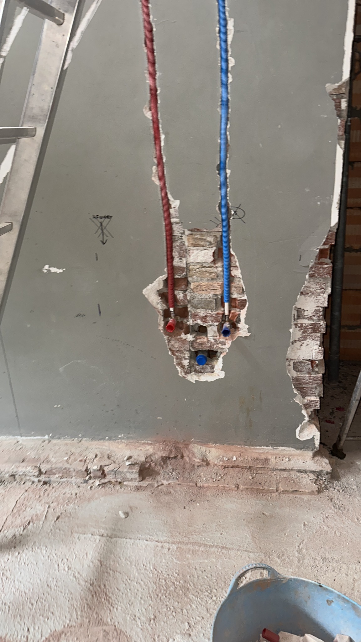 Wykute w ścianie bruzdy z wystającymi rurami instalacji wodnej (czerwona i niebieska) w trakcie montażu, widoczne fragmenty cegieł i ślady po kuciu.