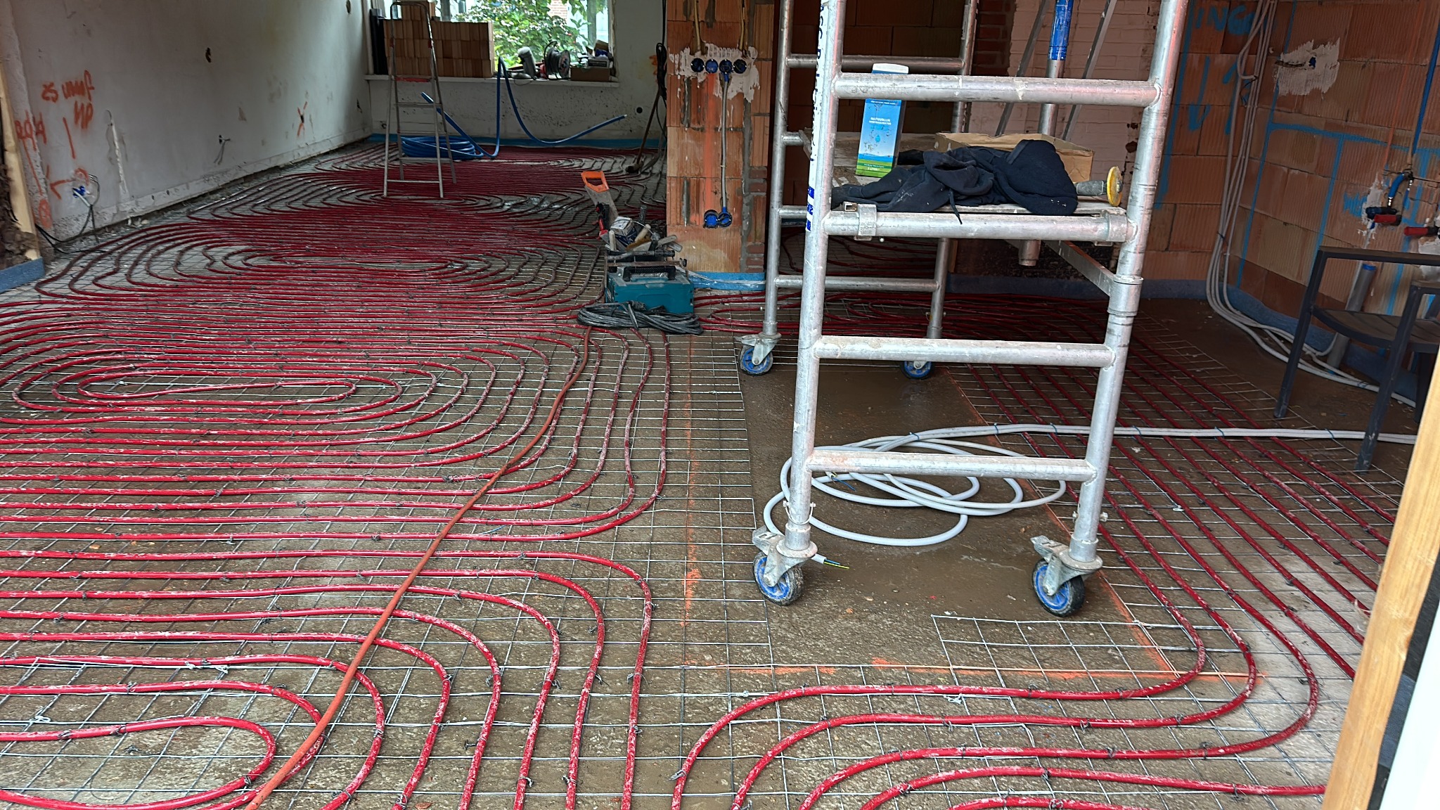 Czerwone rury grzewcze ułożone na metalowej siatce w trakcie montażu ogrzewania podłogowego, widoczny fragment drabiny i narzędzia budowlane.