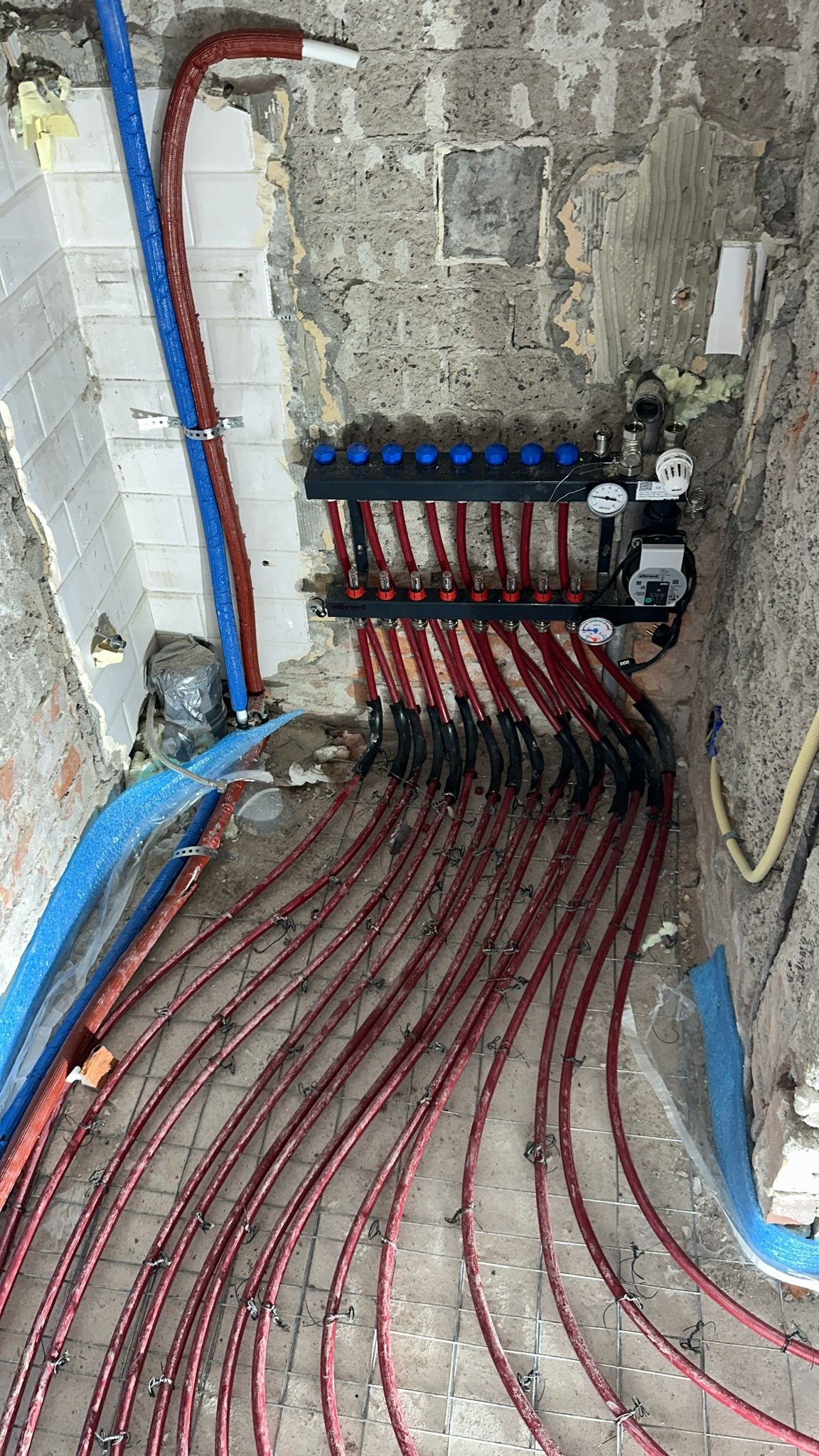 Rozdzielacz ogrzewania podłogowego z czerwonymi rurami ułożonymi na siatce w surowym pomieszczeniu z odsłoniętą cegłą i fragmentami płytek na ścianach, widoczne niebieskie rury i izolacja.