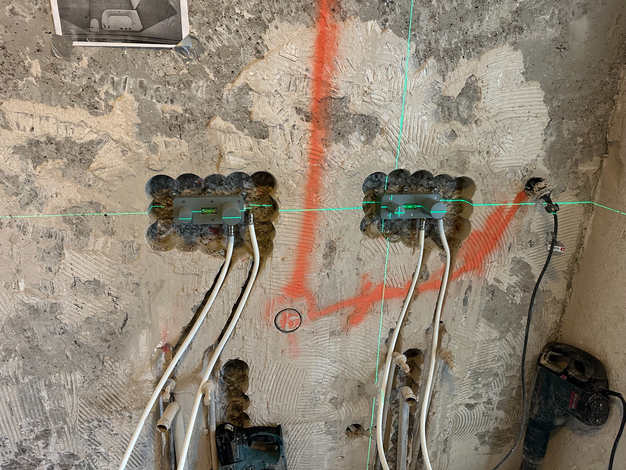 Przygotowanie ściany pod instalację wodno-kanalizacyjną z użyciem poziomicy laserowej, widoczne wycięte otwory, białe rury i ślady po kuciu w surowej ścianie.