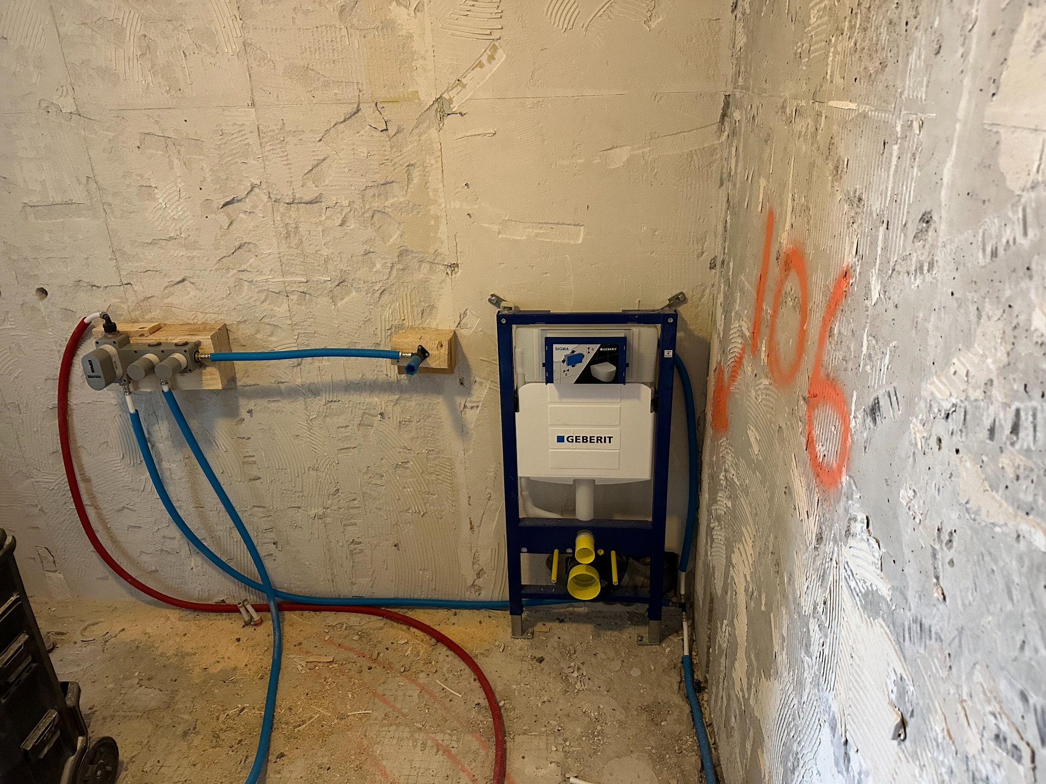 Stelaż podtynkowy Geberit zamontowany na surowej ścianie podczas prac instalacyjnych, widoczne podłączenia wodne i kanalizacyjne, czerwona rura na podłodze, ściana z resztkami zaprawy.