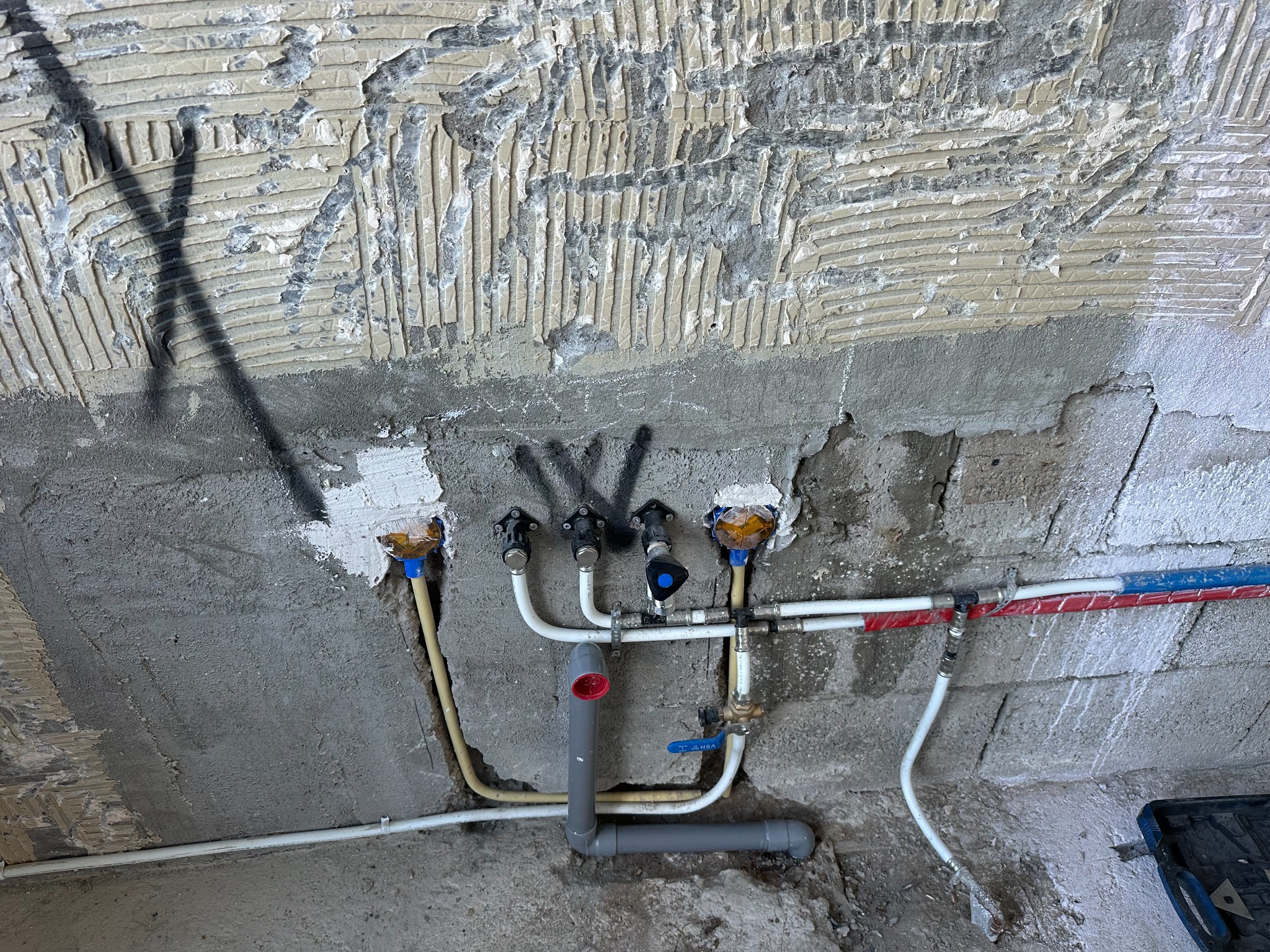 Surowy montaż instalacji wodno-kanalizacyjnej na tle ściany z odsłoniętą strukturą kleju do płytek, widoczne rury PCV, zawory kulowe i złączki, instalacja w trakcie prac, zarys narzędzi w tle.