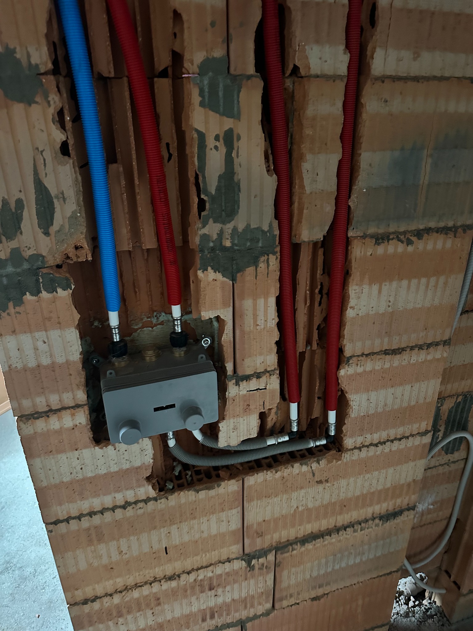 Wnęka w ceglanej ścianie z wystającymi rurami w kolorze niebieskim i czerwonym oraz szarą skrzynką z zaworami, widoczne fragmenty instalacji wodnej i kanalizacyjnej.