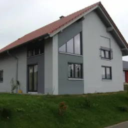 Nowoczesny, dwukondygnacyjny dom z szarą elewacją, ciemnoszarą wstawką z oknami o nietypowym kształcie i roletą zewnętrzną na jednym z okien, z zielonym trawnikiem z przodu.