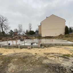 AVA Complex - Wylany fundament z wystającymi prętami zbrojeniowymi, drewnianym szalunkiem i deskami na obrzeżach placu budowy. W tle budynek mieszkalny i pochmurne niebo.