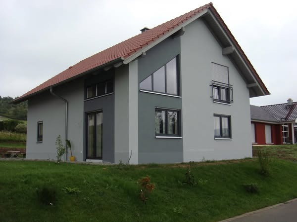 Nowoczesny, dwukondygnacyjny dom z szarą elewacją, ciemnoszarą wstawką z oknami o nietypowym kształcie i roletą zewnętrzną na jednym z okien, z zielonym trawnikiem z przodu.