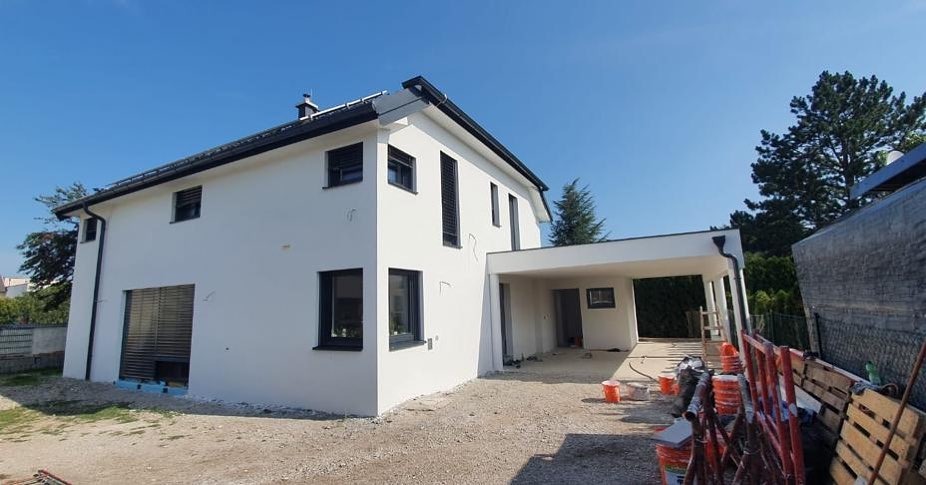 Dwukondygnacyjny dom z białym tynkiem i czarnymi oknami, z dobudowanym garażem, widoczne materiały budowlane i wiaderka z farbą na podwórku.