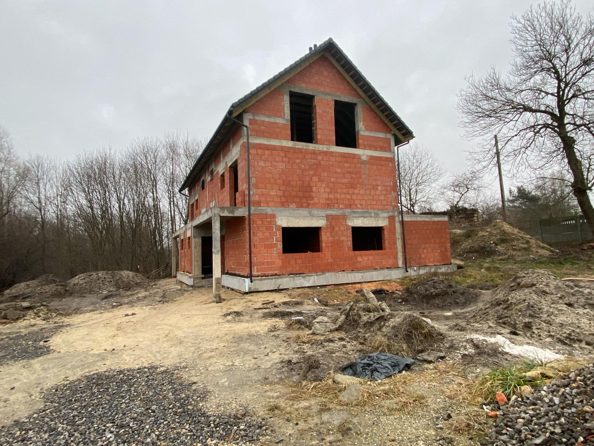 Surowy, dwupiętrowy budynek z czerwonej cegły w trakcie budowy, widoczne puste otwory okienne, szare niebo i otaczający teren z gruzem i piaskiem.