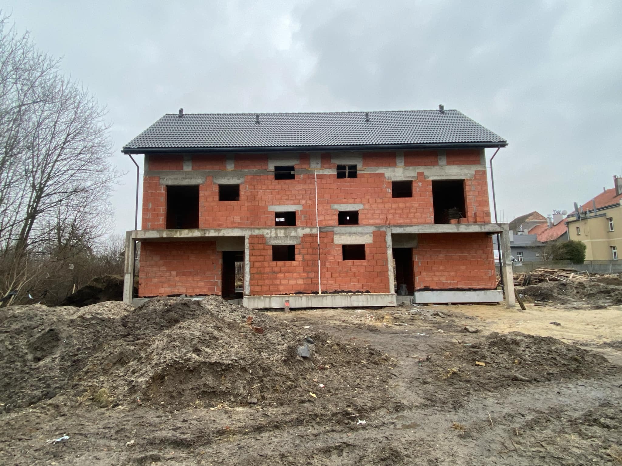 Surowy stan bliźniaka z czerwonej cegły z szarą dachówką, widoczny fundament i stosy ziemi na placu budowy w pochmurny dzień.