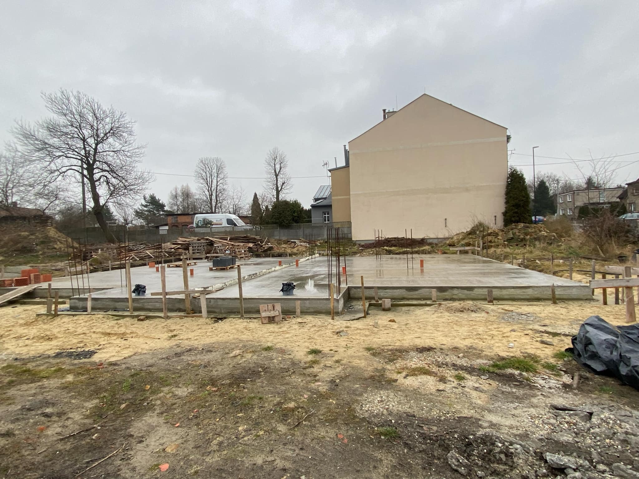 Wylany fundament z wystającymi prętami zbrojeniowymi, drewnianym szalunkiem i deskami na obrzeżach placu budowy. W tle budynek mieszkalny i pochmurne niebo.