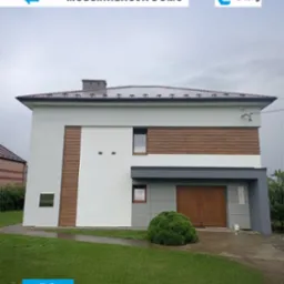 Zdjęcie domu po modernizacji, z nową elewacją w odcieniach bieli, szarości i drewna. Widoczny fragment trawnika i logo firmy. Ujęcie w pochmurny dzień.