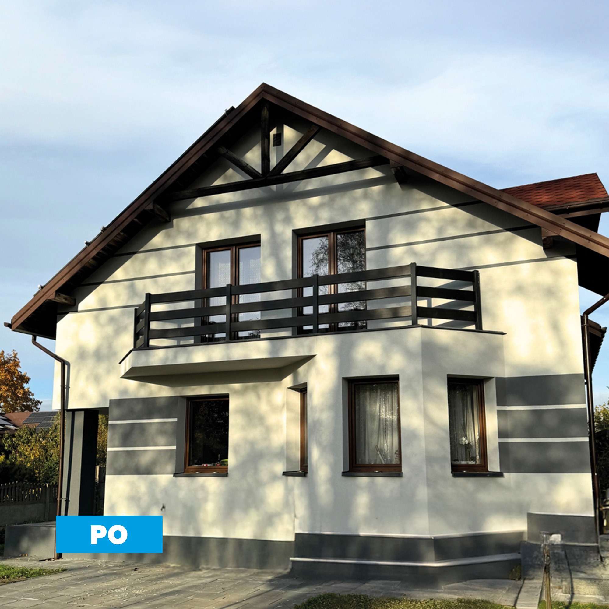 Elewacja domu po ociepleniu, z widocznymi oknami w brązowych ramach i balkonem z ciemną balustradą. Na ścianie widoczny napis 'PO'.