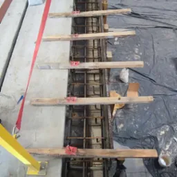 Zbrojenie pionowe w szalunku, zabezpieczone drewnianymi rozpórkami i czerwonymi zaciskami, widoczne elementy rusztowania i folia ochronna.