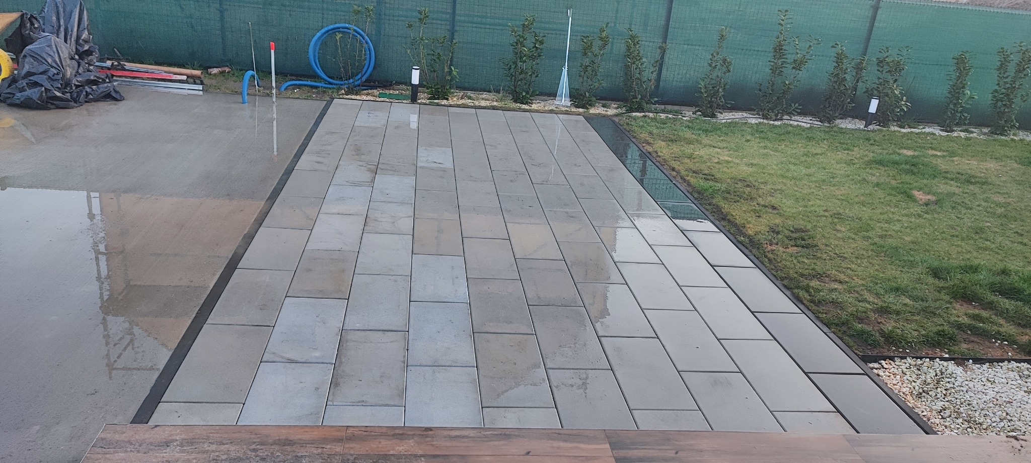 Ułożona, wilgotna nawierzchnia z prostokątnych płyt betonowych na zewnątrz, z widocznym trawnikiem i elementami zieleni w tle.