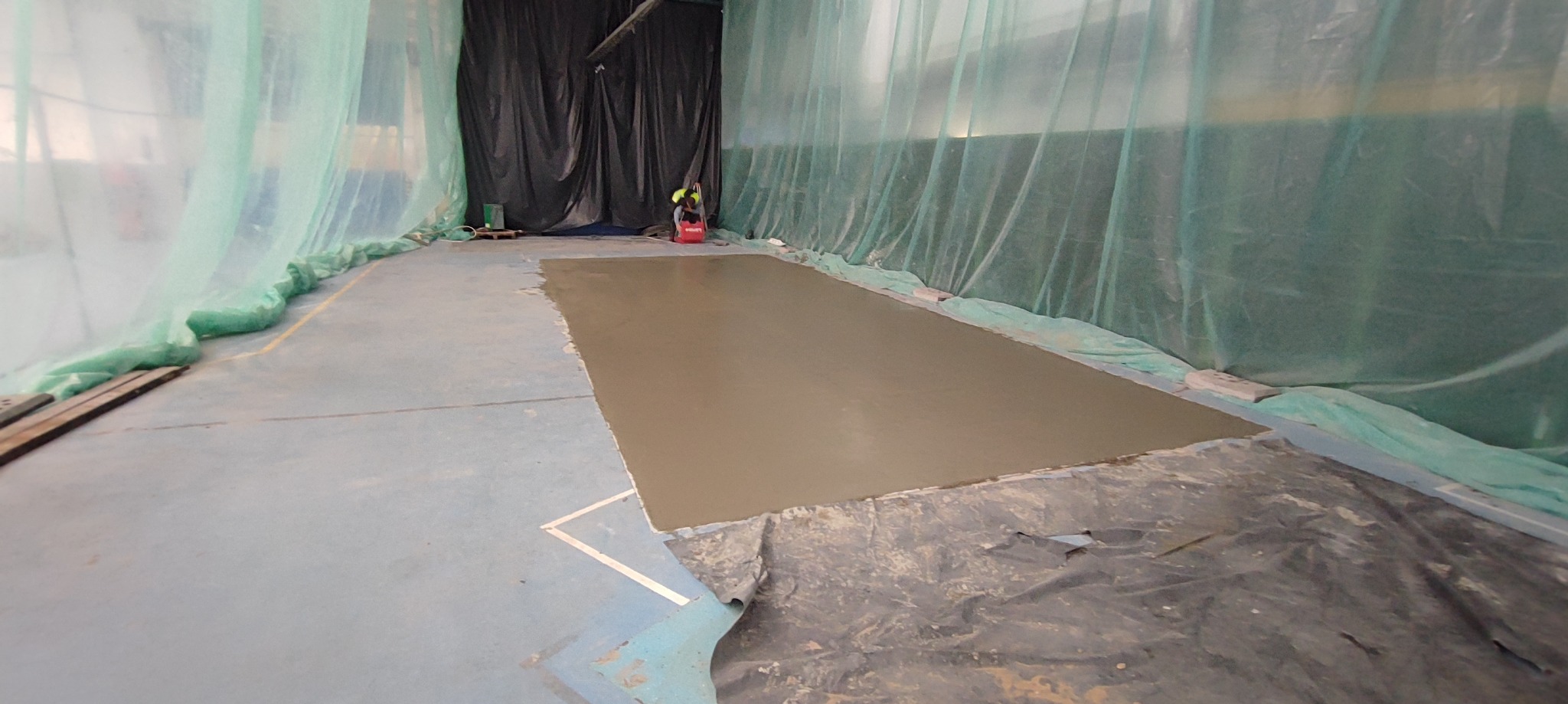 Wyrównywanie posadzki w hali sportowej z użyciem wylewki samopoziomującej, zabezpieczonej folią i kurtyną ochronną.
