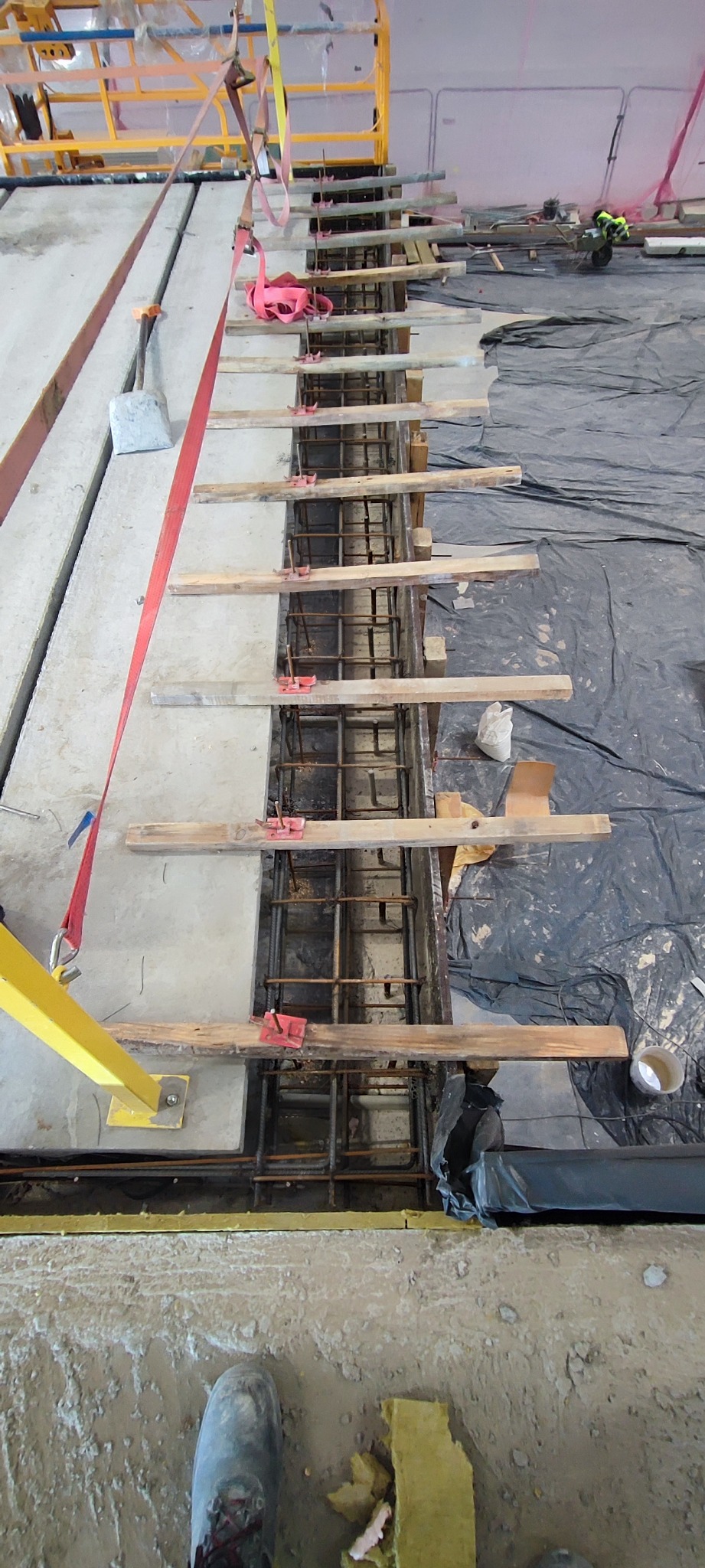 Zbrojenie pionowe w szalunku, zabezpieczone drewnianymi rozpórkami i czerwonymi zaciskami, widoczne elementy rusztowania i folia ochronna.